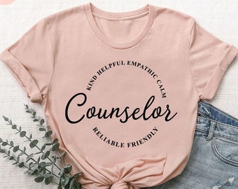 May include: A heather peach t-shirt with the word "Counselor" in a black script font, encircled by the words "Kind, Helpful, Empathetic, Calm, Reliable, Friendly." The shirt is styled with denim shorts and a sprig of greenery.