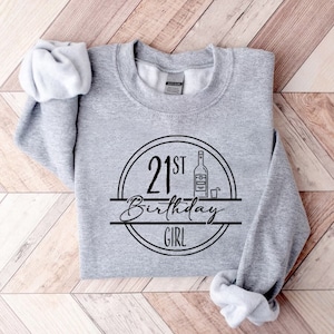 K&ouml;nnte beinhalten: Hellgraues Sweatshirt mit schwarzem Aufdruck "21st Birthday Girl" in einem Kreis, mit einer Flaschen- und Schnapsglas-Illustration. Das Sweatshirt liegt gefaltet auf einer hellbraunen Holzoberfl&auml;che.