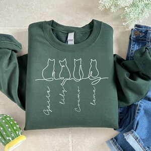 May include: A dark green sweatshirt with a white outline of four cats sitting in a row. The cats are labeled "Gracie", "Lily", "Cosmo", and "Luna".
