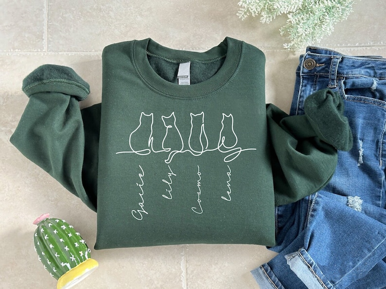 May include: A dark green sweatshirt with a white outline of four cats sitting in a row. The cats are labeled "Gracie", "Lily", "Cosmo", and "Luna".