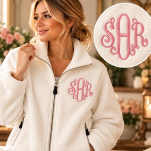 May include: A white fleece zip-up jacket featuring a high collar and long sleeves. The front of the jacket is embroidered with a pink monogram featuring the letters SAR in a decorative script font. The jacket includes a silver metal zipper.