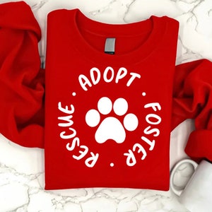 May include: A bright red sweatshirt with the words "Adopt. Rescue. Foster" encircling a white paw print. The long-sleeved top is folded, and a white mug is visible in the lower right corner. The design promotes pet adoption.