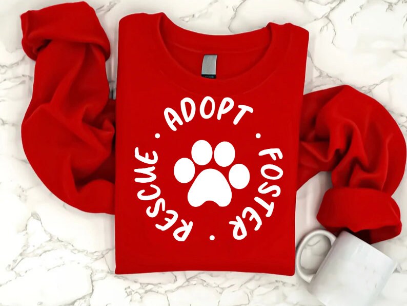 May include: A bright red sweatshirt with the words "Adopt. Rescue. Foster" encircling a white paw print. The long-sleeved top is folded, and a white mug is visible in the lower right corner. The design promotes pet adoption.