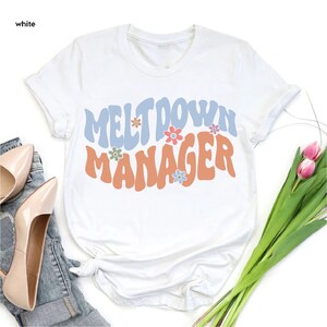 May include: White t-shirt with a retro-style graphic that says "Meltdown Manager" in blue and orange letters with colorful flowers.