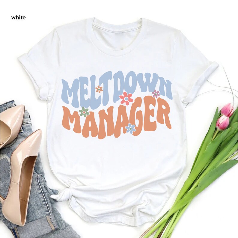 May include: White t-shirt with a retro-style graphic that says "Meltdown Manager" in blue and orange letters with colorful flowers.