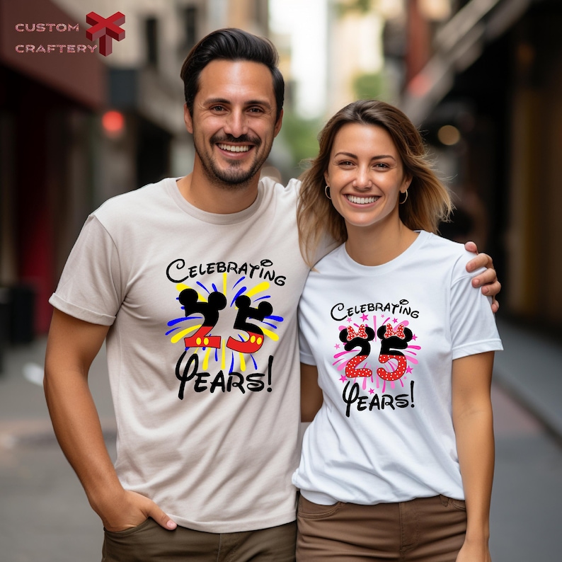May include: Two white t-shirts with a graphic design of Mickey Mouse ears and fireworks. The text on the shirts reads "Celebrating 25 Years!"