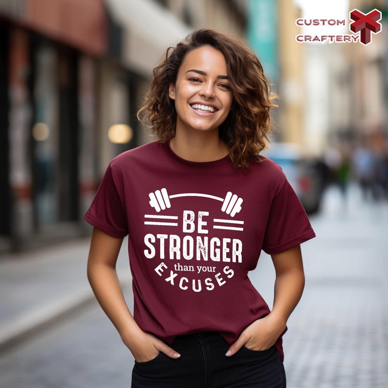 May include: A maroon t-shirt with the text "BE STRONGER than your EXCUSES" in white, along with a barbell graphic. The shirt is being worn by a person with hands in their pockets, smiling in front of a blurred background.