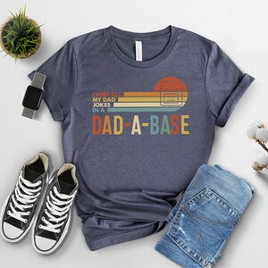 May include: Navy blue t-shirt with the text "I keep all my dad jokes in a DAD-A-BASE" in a retro design. The graphic features a sunset-colored rectangle and a computer server. The shirt is paired with black sneakers and blue jeans.