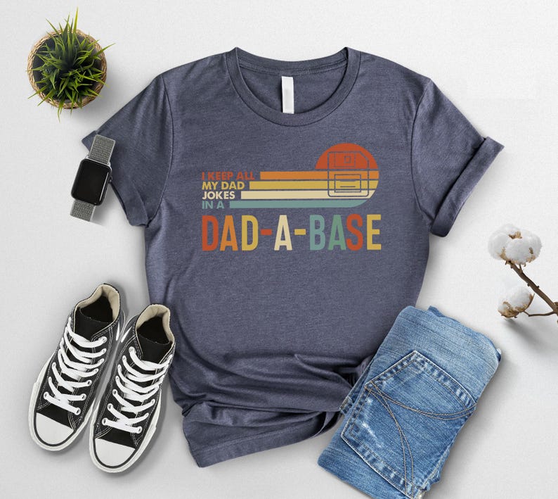 May include: Navy blue t-shirt with the text "I keep all my dad jokes in a DAD-A-BASE" in a retro design. The graphic features a sunset-colored rectangle and a computer server. The shirt is paired with black sneakers and blue jeans.