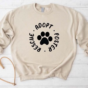 May include: A beige sweatshirt with a black paw print and the words "Adopt, Rescue, Foster" in a circular design. The sweatshirt has long sleeves and a crew neck. The background is a light wood-grain pattern.