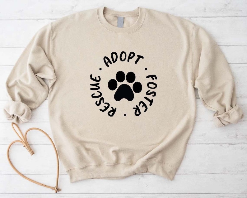 May include: A beige sweatshirt with a black paw print and the words "Adopt, Rescue, Foster" in a circular design. The sweatshirt has long sleeves and a crew neck. The background is a light wood-grain pattern.
