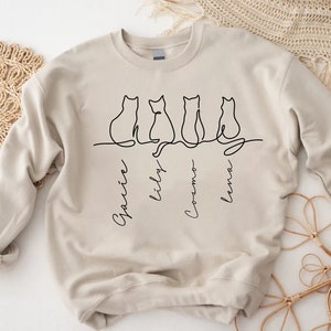 May include: A beige sweatshirt with a black line drawing of four cats sitting in a row. The cats are labeled "Garvin", "Lily", "Cosmo", and "Lena".