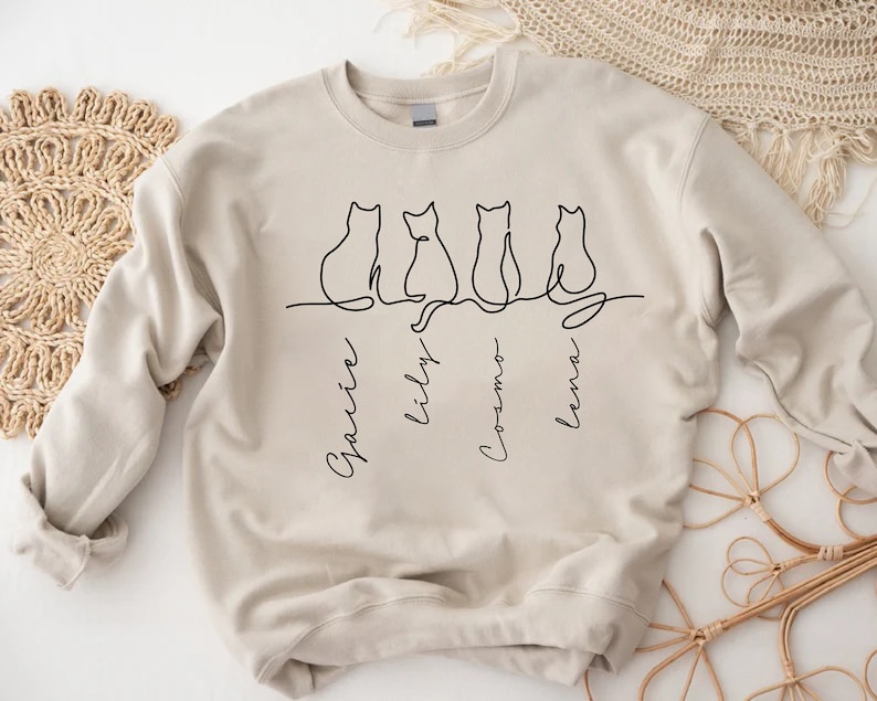 May include: A beige sweatshirt with a black line drawing of four cats sitting in a row. The cats are labeled "Garvin", "Lily", "Cosmo", and "Lena".