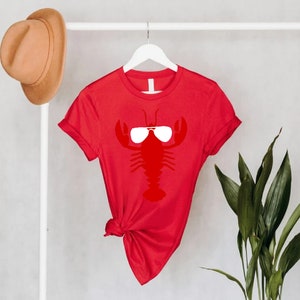 Pu&ograve; includere: Una maglietta rossa con una grafica bianca di un'aragosta con occhiali da sole. La maglietta &egrave; appesa a un gancio bianco. Un cappello marrone &egrave; appeso sopra. Piante verdi sullo sfondo.