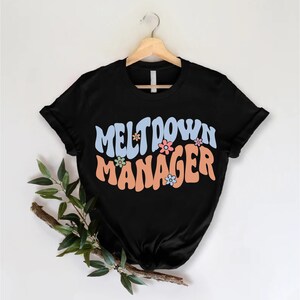 May include: Black t-shirt with the text "Meltdown Manager" in a retro font with colorful flowers.