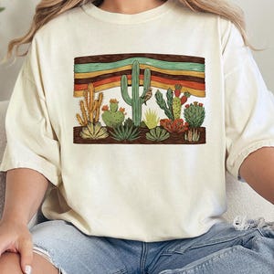 K&ouml;nnte beinhalten: Cremefarbenes T-Shirt mit einer W&uuml;stenlandschaft mit verschiedenen Kakteen und Sukkulenten. Die Grafik enth&auml;lt ein horizontales Band mit Streifen in Braun, Orange, Gelb und Gr&uuml;n. Das Shirt hat eine lockere Passform.
