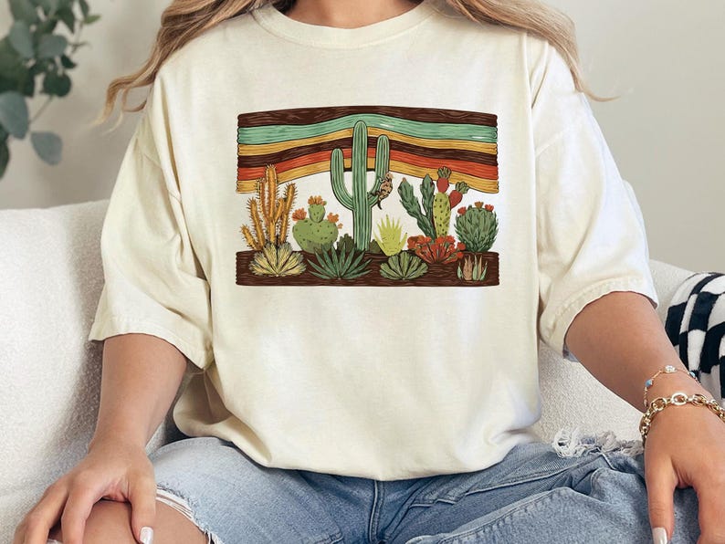 K&ouml;nnte beinhalten: Cremefarbenes T-Shirt mit einer W&uuml;stenlandschaft mit verschiedenen Kakteen und Sukkulenten. Die Grafik enth&auml;lt ein horizontales Band mit Streifen in Braun, Orange, Gelb und Gr&uuml;n. Das Shirt hat eine lockere Passform.