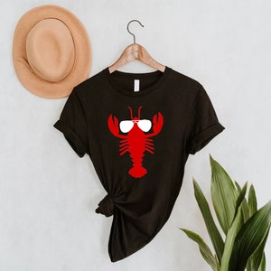 Pu&ograve; includere: T-shirt nero con un'aragosta rossa con occhiali da sole bianchi. Il disegno dell'aragosta &egrave; centrato sulla maglietta. Un cappello marrone &egrave; appeso al muro dietro la maglietta e a destra si vedono foglie verdi.