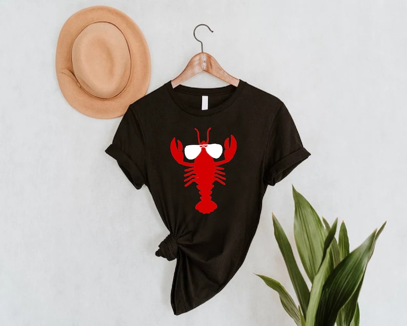 Pu&ograve; includere: T-shirt nero con un'aragosta rossa con occhiali da sole bianchi. Il disegno dell'aragosta &egrave; centrato sulla maglietta. Un cappello marrone &egrave; appeso al muro dietro la maglietta e a destra si vedono foglie verdi.