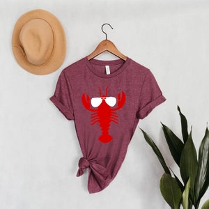 Pu&ograve; includere: T-shirt bordeaux con grafica di aragosta rossa con occhiali da sole. La maglietta &egrave; appesa a una gruccia di legno contro un muro bianco. Un cappello beige &egrave; sul muro a sinistra e piante verdi a destra.