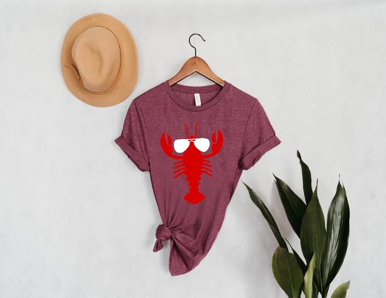 Pu&ograve; includere: T-shirt bordeaux con grafica di aragosta rossa con occhiali da sole. La maglietta &egrave; appesa a una gruccia di legno contro un muro bianco. Un cappello beige &egrave; sul muro a sinistra e piante verdi a destra.
