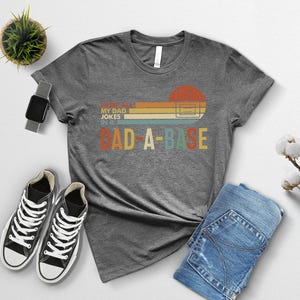 May include: A heather gray t-shirt with the text "I love my dad jokes in a dad-a-base" in a retro design. The graphic includes a sunset-colored rectangle and a computer server icon. The shirt is paired with black sneakers and blue jeans.