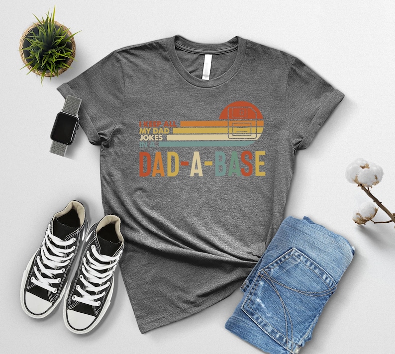May include: A heather gray t-shirt with the text "I love my dad jokes in a dad-a-base" in a retro design. The graphic includes a sunset-colored rectangle and a computer server icon. The shirt is paired with black sneakers and blue jeans.