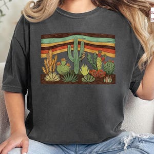 K&ouml;nnte beinhalten: Dunkelgraues T-Shirt mit einem W&uuml;stenkaktus-Design. Die Grafik zeigt verschiedene Kakteen und einen gestreiften Hintergrund in Orange, Gelb und Gr&uuml;n. Das Shirt ist ein bequemer, l&auml;ssiger Stil.