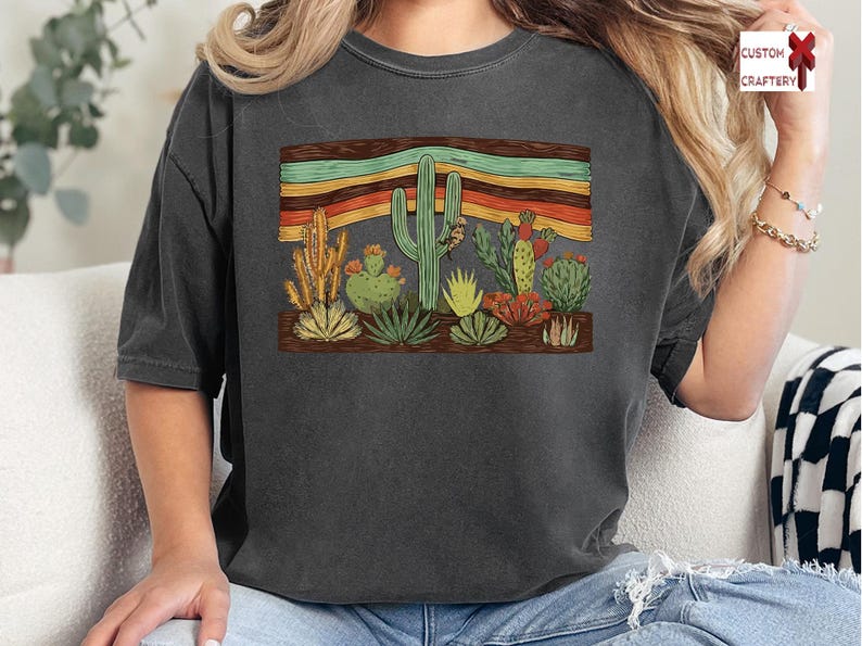 K&ouml;nnte beinhalten: Dunkelgraues T-Shirt mit einem W&uuml;stenkaktus-Design. Die Grafik zeigt verschiedene Kakteen und einen gestreiften Hintergrund in Orange, Gelb und Gr&uuml;n. Das Shirt ist ein bequemer, l&auml;ssiger Stil.