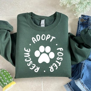 May include: A dark green sweatshirt with the words "ADOPT. RESCUE. FOSTER." encircling a white paw print. The sweatshirt is folded next to a pair of blue jeans.