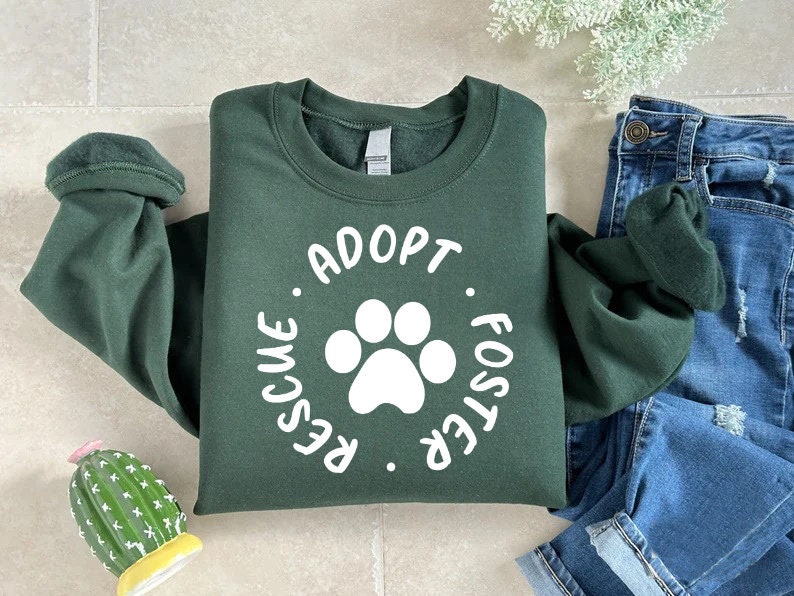 May include: A dark green sweatshirt with the words "ADOPT. RESCUE. FOSTER." encircling a white paw print. The sweatshirt is folded next to a pair of blue jeans.