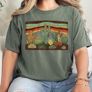 K&ouml;nnte beinhalten: Olivgr&uuml;nes T-Shirt mit einem W&uuml;stenlandschaftsdesign. Die Grafik zeigt verschiedene Kakteen und Sukkulenten in Gr&uuml;n-, Gelb- und Rott&ouml;nen, vor einem Hintergrund aus horizontalen Streifen in Orange, Gelb und T&uuml;rkis.