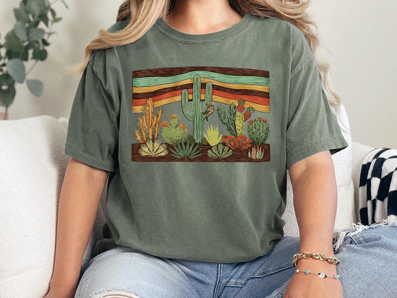 K&ouml;nnte beinhalten: Olivgr&uuml;nes T-Shirt mit einem W&uuml;stenlandschaftsdesign. Die Grafik zeigt verschiedene Kakteen und Sukkulenten in Gr&uuml;n-, Gelb- und Rott&ouml;nen, vor einem Hintergrund aus horizontalen Streifen in Orange, Gelb und T&uuml;rkis.