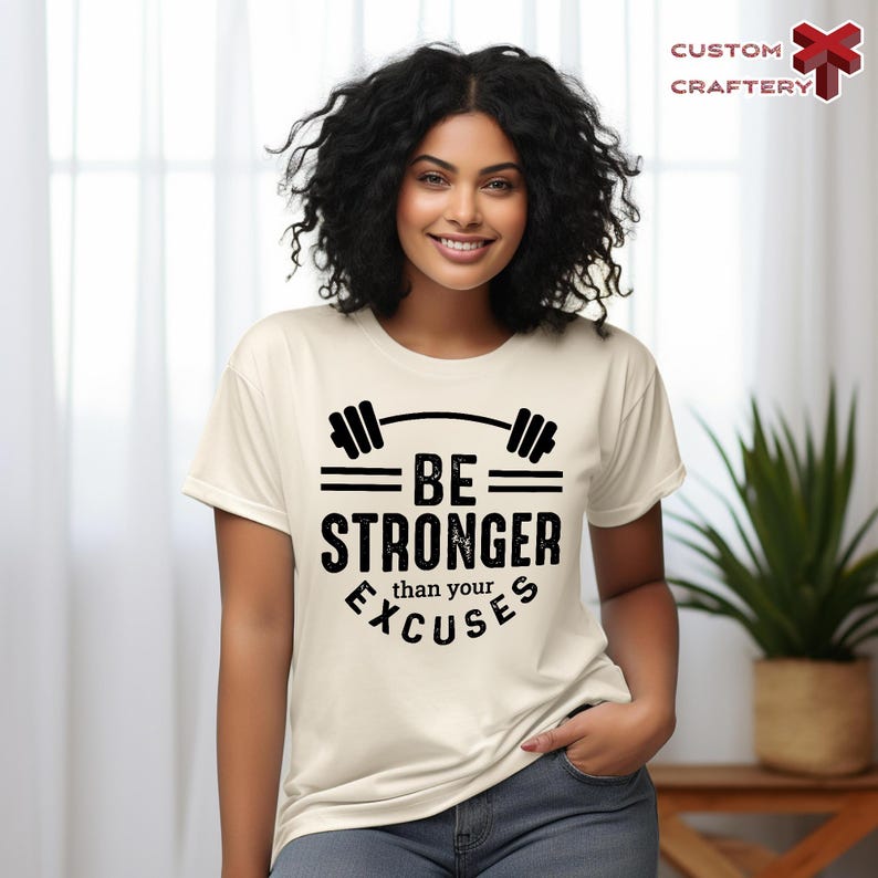 May include: Cream-colored t-shirt with a black barbell graphic and the text "BE STRONGER than your EXCUSES." The shirt is made of a soft material and is suitable for workouts or casual wear. The Custom Craftery logo is in the top right corner.