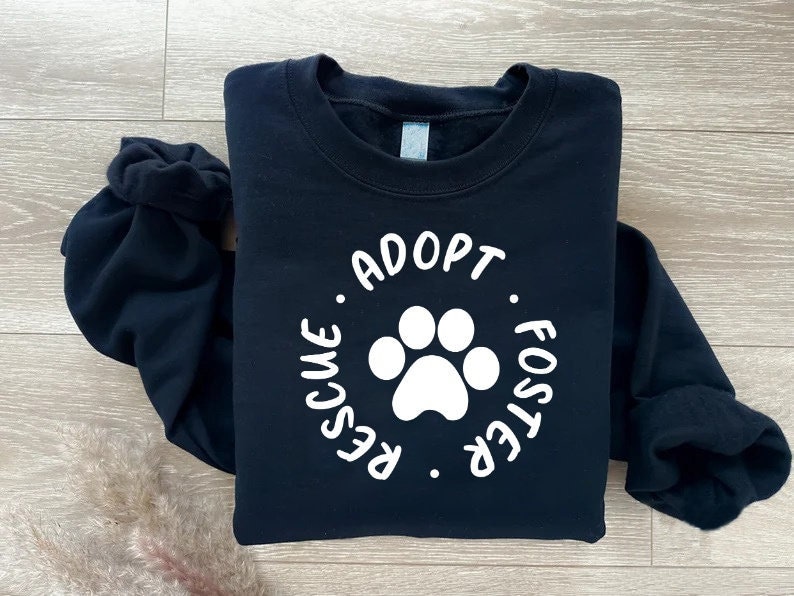 May include: A navy blue sweatshirt with the words "RESCUE. ADOPT. FOSTER." encircling a white paw print. The sweatshirt is folded, with the sleeves bunched up. The background is a light wood grain.