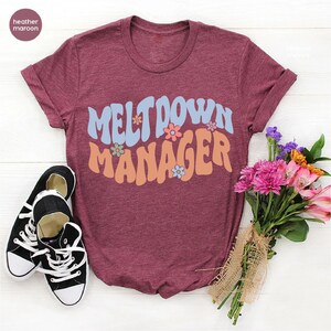 May include: Heather maroon t-shirt with the text "Meltdown Manager" in a retro font with colorful flowers.