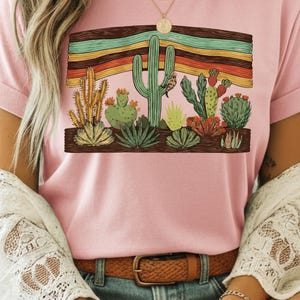 K&ouml;nnte beinhalten: Hellrosa T-Shirt mit einem W&uuml;stenmotiv. Das Design zeigt verschiedene Kakteen und Sukkulenten, vor einem Hintergrund aus bunten horizontalen Streifen in einem Holzrahmen. Das T-Shirt hat umgekrempelte &Auml;rmel.