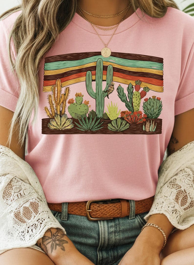 K&ouml;nnte beinhalten: Hellrosa T-Shirt mit einem W&uuml;stenmotiv. Das Design zeigt verschiedene Kakteen und Sukkulenten, vor einem Hintergrund aus bunten horizontalen Streifen in einem Holzrahmen. Das T-Shirt hat umgekrempelte &Auml;rmel.