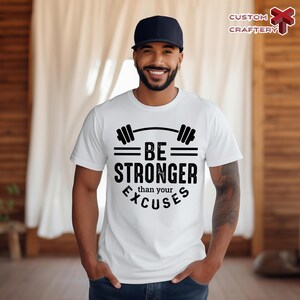 May include: White t-shirt with a black graphic that reads "BE STRONGER than your EXCUSES" with a barbell design above the words. The shirt is worn by a person wearing a black baseball cap.