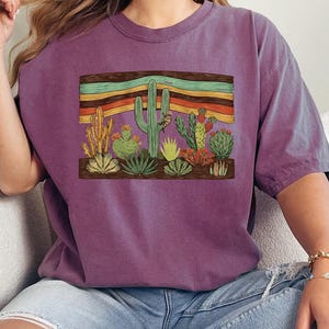 K&ouml;nnte beinhalten: Ein lila T-Shirt mit einer W&uuml;stenszene mit verschiedenen Kakteen und Sukkulenten. Die Grafik enth&auml;lt einen Holzrahmen mit bunten horizontalen Streifen &uuml;ber den Pflanzen. Das Shirt ist aus weichem Material.
