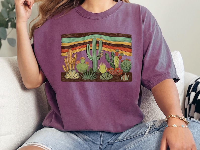 K&ouml;nnte beinhalten: Ein lila T-Shirt mit einer W&uuml;stenszene mit verschiedenen Kakteen und Sukkulenten. Die Grafik enth&auml;lt einen Holzrahmen mit bunten horizontalen Streifen &uuml;ber den Pflanzen. Das Shirt ist aus weichem Material.