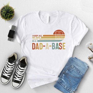 May include: White t-shirt with a retro graphic that reads "I keep all my dad jokes in a DAD-A-BASE." The design features a floppy disk icon and horizontal stripes in orange, yellow, and brown. The shirt is paired with blue jeans and black sneakers.