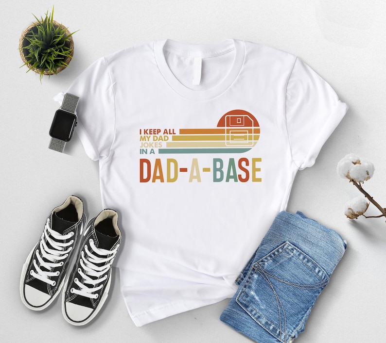 May include: White t-shirt with a retro graphic that reads "I keep all my dad jokes in a DAD-A-BASE." The design features a floppy disk icon and horizontal stripes in orange, yellow, and brown. The shirt is paired with blue jeans and black sneakers.