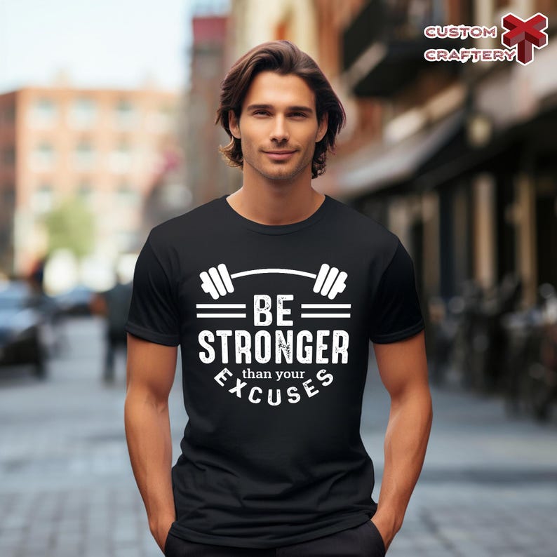 May include: Black t-shirt with the text "BE STRONGER than your EXCUSES" in white. The design includes a barbell graphic above the text. The shirt is worn by a person in an outdoor setting.