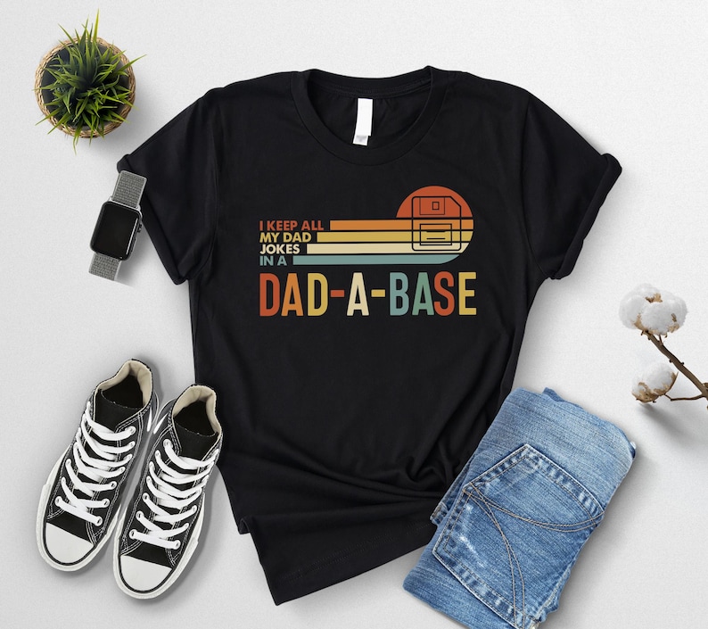 May include: Black t-shirt with the text "I KEEP ALL MY DAD JOKES IN A DAD-A-BASE" in a retro design. The graphic includes a floppy disk and horizontal stripes in orange, yellow, and blue. The shirt is paired with sneakers and jeans.