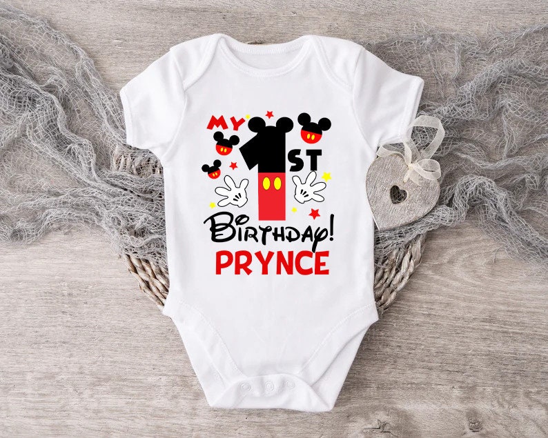 Puede incluir: Un body blanco para beb&eacute; con un dise&ntilde;o de Mickey Mouse rojo y negro. El body dice "My 1st Birthday! PRYNCE".