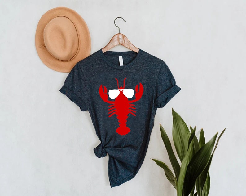 Pu&ograve; includere: Una maglietta grigio scuro con un'aragosta rossa con occhiali da sole bianchi. Il disegno dell'aragosta &egrave; centrato sulla maglietta. Un cappello marrone &egrave; appeso al muro dietro la maglietta.