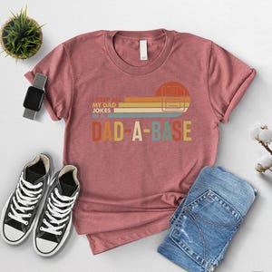 May include: A dusty rose t-shirt with the text "I KEEP ALL MY DAD JOKES IN A DAD-A-BASE" in a retro design. The shirt is paired with blue jeans and black and white sneakers. A smartwatch and a small potted plant are also in the image.