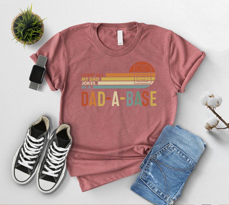May include: A dusty rose t-shirt with the text "I KEEP ALL MY DAD JOKES IN A DAD-A-BASE" in a retro design. The shirt is paired with blue jeans and black and white sneakers. A smartwatch and a small potted plant are also in the image.