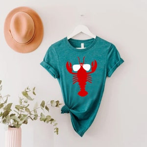 Pu&ograve; includere: T-shirt color turchese con un'aragosta rossa con occhiali da sole bianchi. La maglietta &egrave; appesa a un gancio bianco. Un cappello marrone chiaro &egrave; sulla parete a sinistra della maglietta. Un vaso con vegetazione &egrave; nell'angolo in basso a sinistra.
